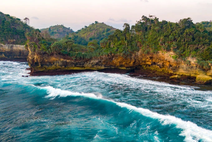 Küstenlandschaft bei Pantai Batu Bengkung in Süd-Malang, Ost-Java, mit felsigen Klippen, dichter Vegetation und starkem Wellengang
